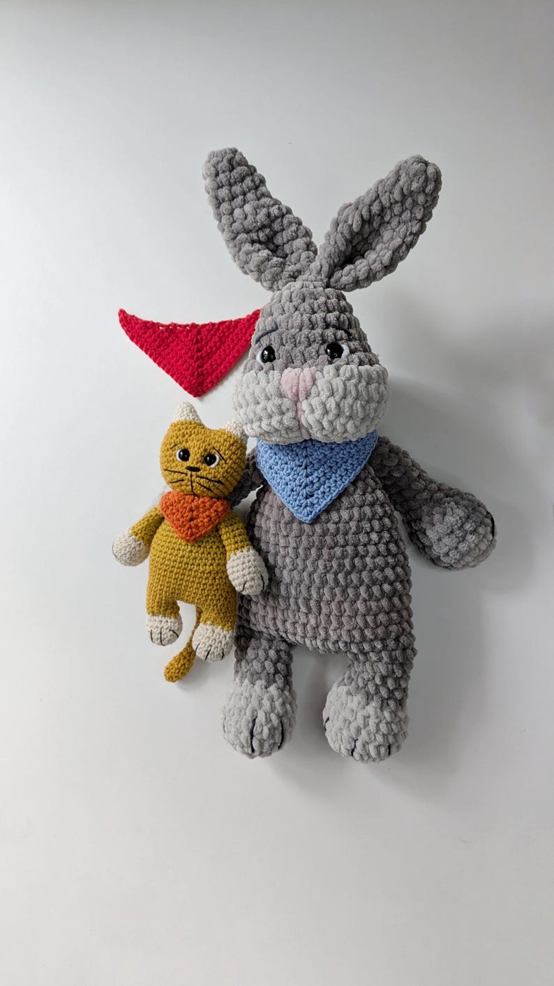 May include: A large gray crocheted rabbit with long ears and a light blue bandana, a smaller yellow crocheted cat with an orange bandana, and a separate red triangular bandana. The rabbit has a pink nose and black eyes, while the cat has black eyes.