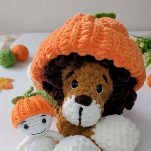 May include: A plush lion toy with dark brown mane and white muzzle wearing a large orange pumpkin hat, positioned next to a small white crochet doll wearing a matching orange pumpkin hat with a green stem, crafted from soft yarn.