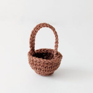 May include: A small, brown crochet basket with a handle. The basket is round with a textured appearance, and the handle is arched. The basket is likely handmade and suitable for small items or decorative purposes.