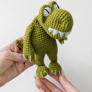 Puede incluir: Un juguete de dinosaurio de ganchillo verde hecho a mano con detalles blancos en los dientes y las garras. El dinosaurio tiene una expresi&oacute;n amigable y se sostiene sobre un fondo blanco. El juguete es un art&iacute;culo lindo y caprichoso.