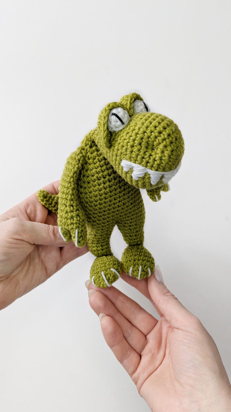Puede incluir: Un juguete de dinosaurio de ganchillo verde hecho a mano con detalles blancos en los dientes y las garras. El dinosaurio tiene una expresi&oacute;n amigable y se sostiene sobre un fondo blanco. El juguete es un art&iacute;culo lindo y caprichoso.