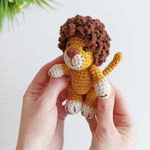 May include: A small crocheted lion toy with a brown mane, yellow body, and white paws. The lion is being held in two hands.