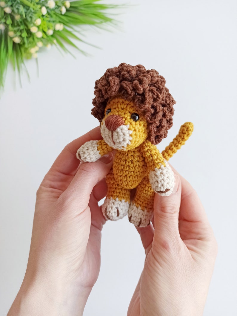 May include: A small crocheted lion toy with a brown mane, yellow body, and white paws. The lion is being held in two hands.