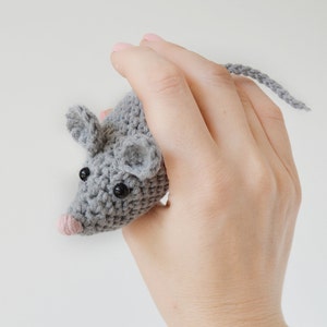May include: A gray crocheted mouse toy with black eyes and a pink nose. The mouse is being held in a hand.