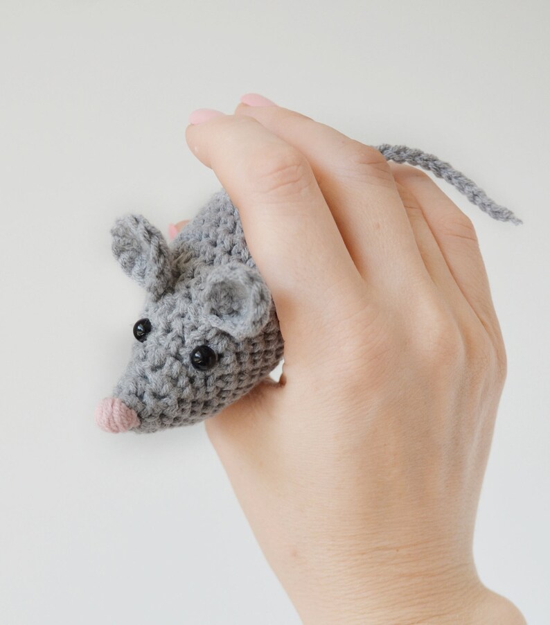May include: A gray crocheted mouse toy with black eyes and a pink nose. The mouse is being held in a hand.