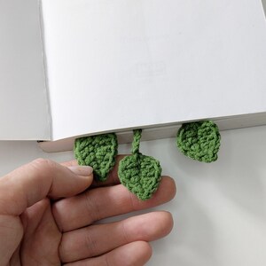 Puede incluir: Un marcap&aacute;ginas de ganchillo con tres peque&ntilde;as hojas verdes texturizadas unidas a un tallo central. Las hojas est&aacute;n hechas de hilo con puntadas visibles, dise&ntilde;adas para colocarse entre las p&aacute;ginas de un libro blanco abierto.