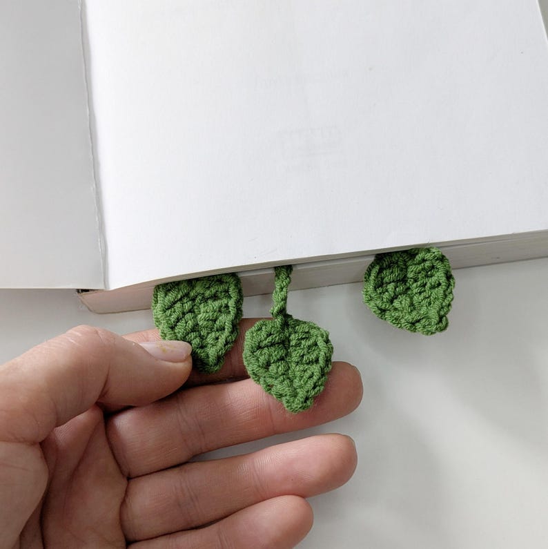 Puede incluir: Un marcap&aacute;ginas de ganchillo con tres peque&ntilde;as hojas verdes texturizadas unidas a un tallo central. Las hojas est&aacute;n hechas de hilo con puntadas visibles, dise&ntilde;adas para colocarse entre las p&aacute;ginas de un libro blanco abierto.