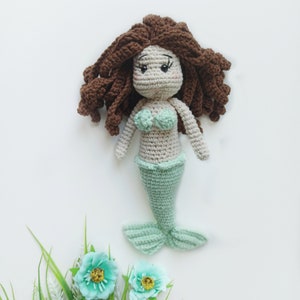 May include: A crocheted mermaid doll with brown hair, a light green tail, and a light green and white top. The doll is sitting on a white background with a green plant with blue flowers.