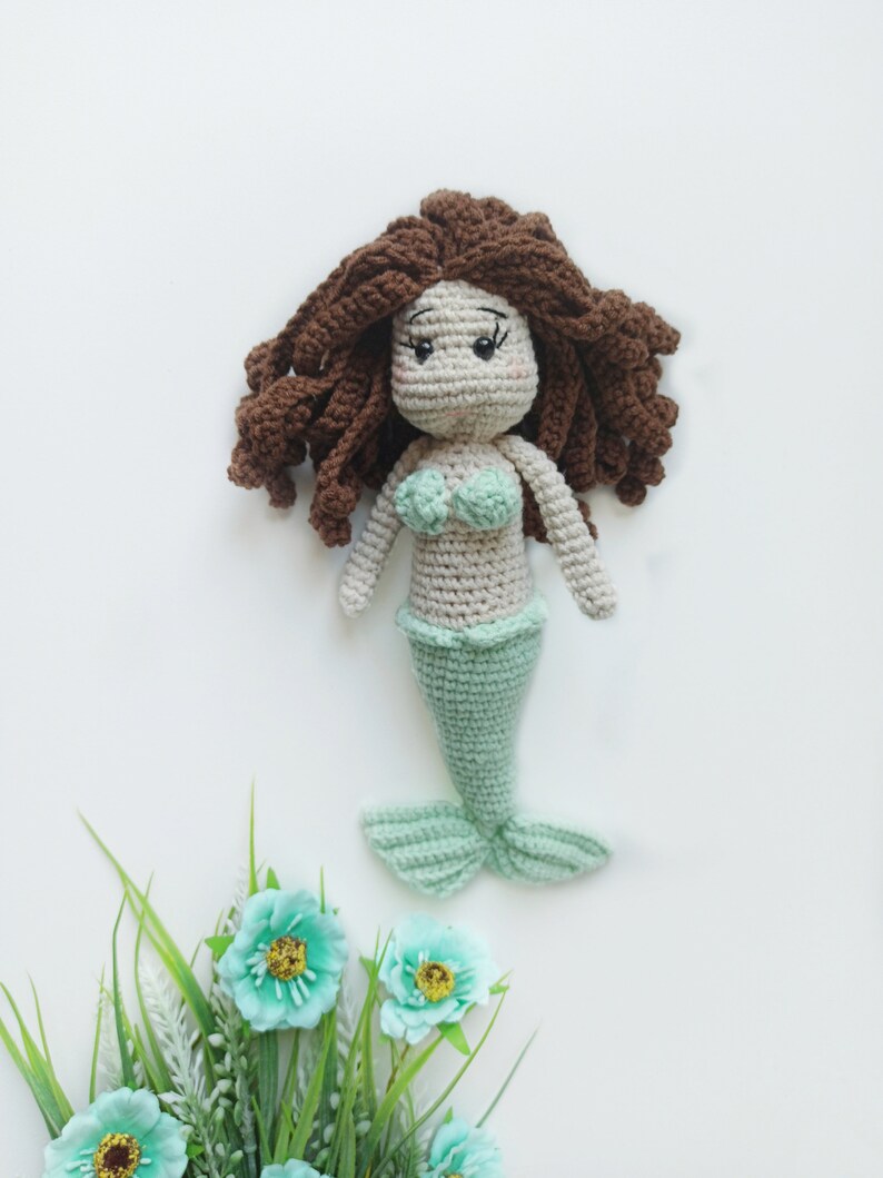 May include: A crocheted mermaid doll with brown hair, a light green tail, and a light green and white top. The doll is sitting on a white background with a green plant with blue flowers.