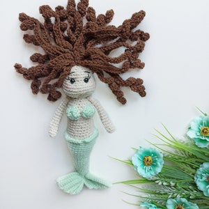 May include: A crocheted mermaid doll with brown hair, a green tail, and a white top. The doll is sitting on a white surface with green plants and blue flowers.