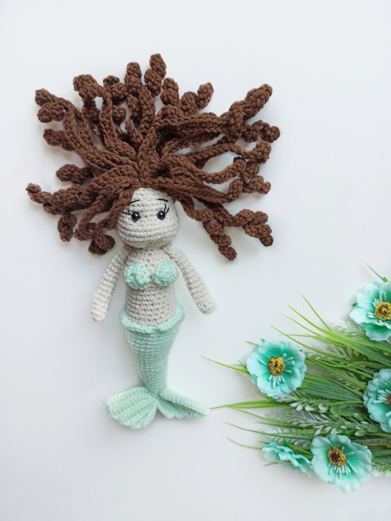 May include: A crocheted mermaid doll with brown hair, a green tail, and a white top. The doll is sitting on a white surface with green plants and blue flowers.