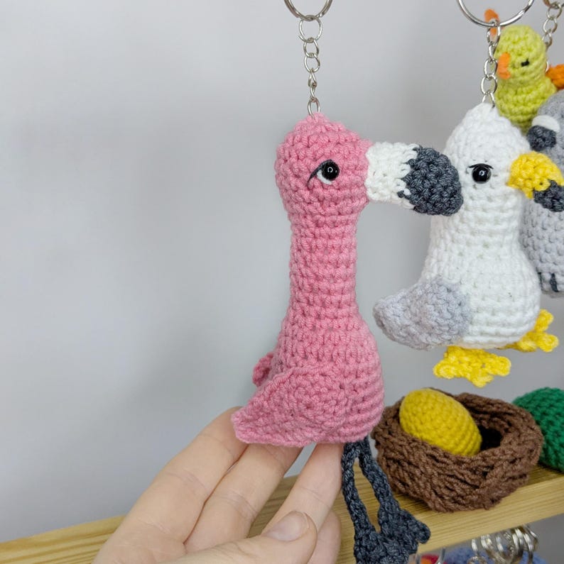 May include: A pink flamingo crochet keychain with a dark gray beak and black eyes, hanging next to a white and gray seagull keychain with a yellow beak. Below them sits a small brown crocheted nest containing a single yellow crocheted egg.