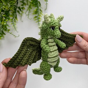 May include: A green crocheted dragon with wings, standing on a white surface.