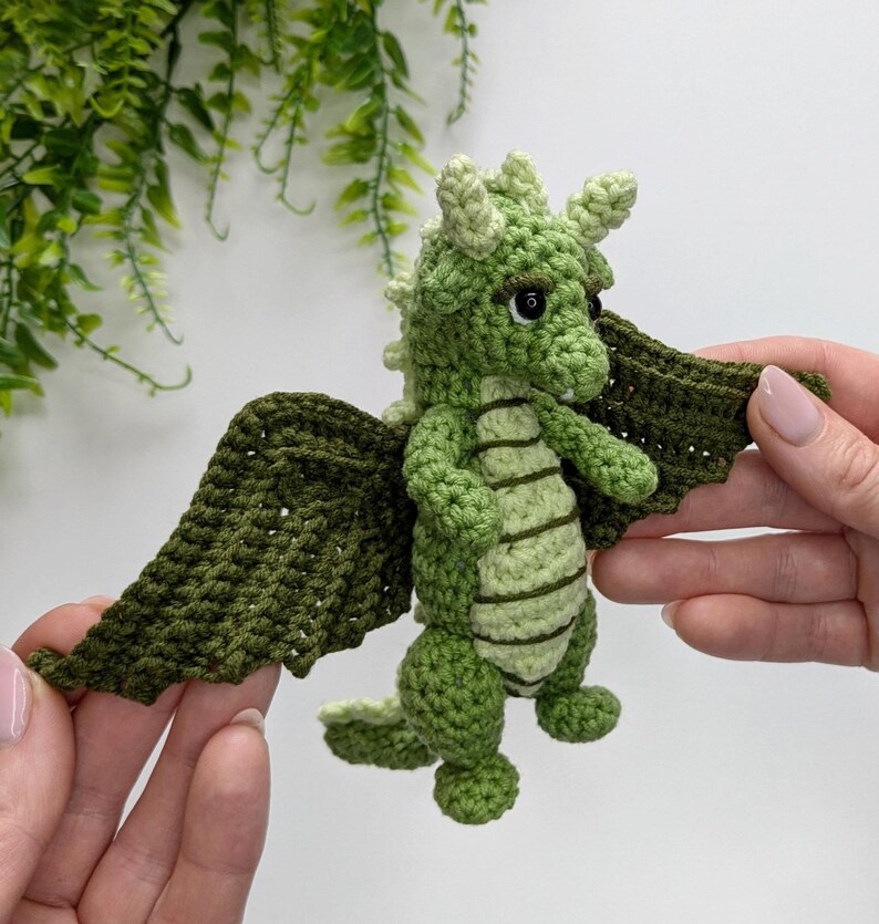 May include: A green crocheted dragon with wings, standing on a white surface.