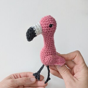 May include: A small, handmade crochet flamingo toy featuring a bright pink body, a long neck, and a curved beak colored in white and dark gray. The flamingo has a single black bead eye and two thin, dark gray crocheted legs with webbed feet.