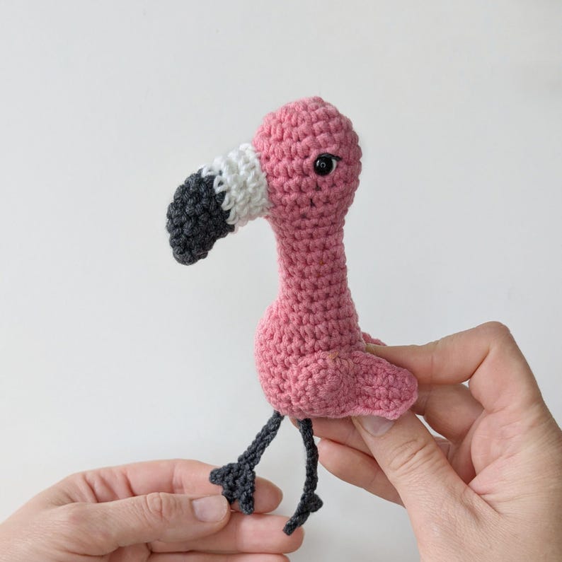 May include: A small, handmade crochet flamingo toy featuring a bright pink body, a long neck, and a curved beak colored in white and dark gray. The flamingo has a single black bead eye and two thin, dark gray crocheted legs with webbed feet.