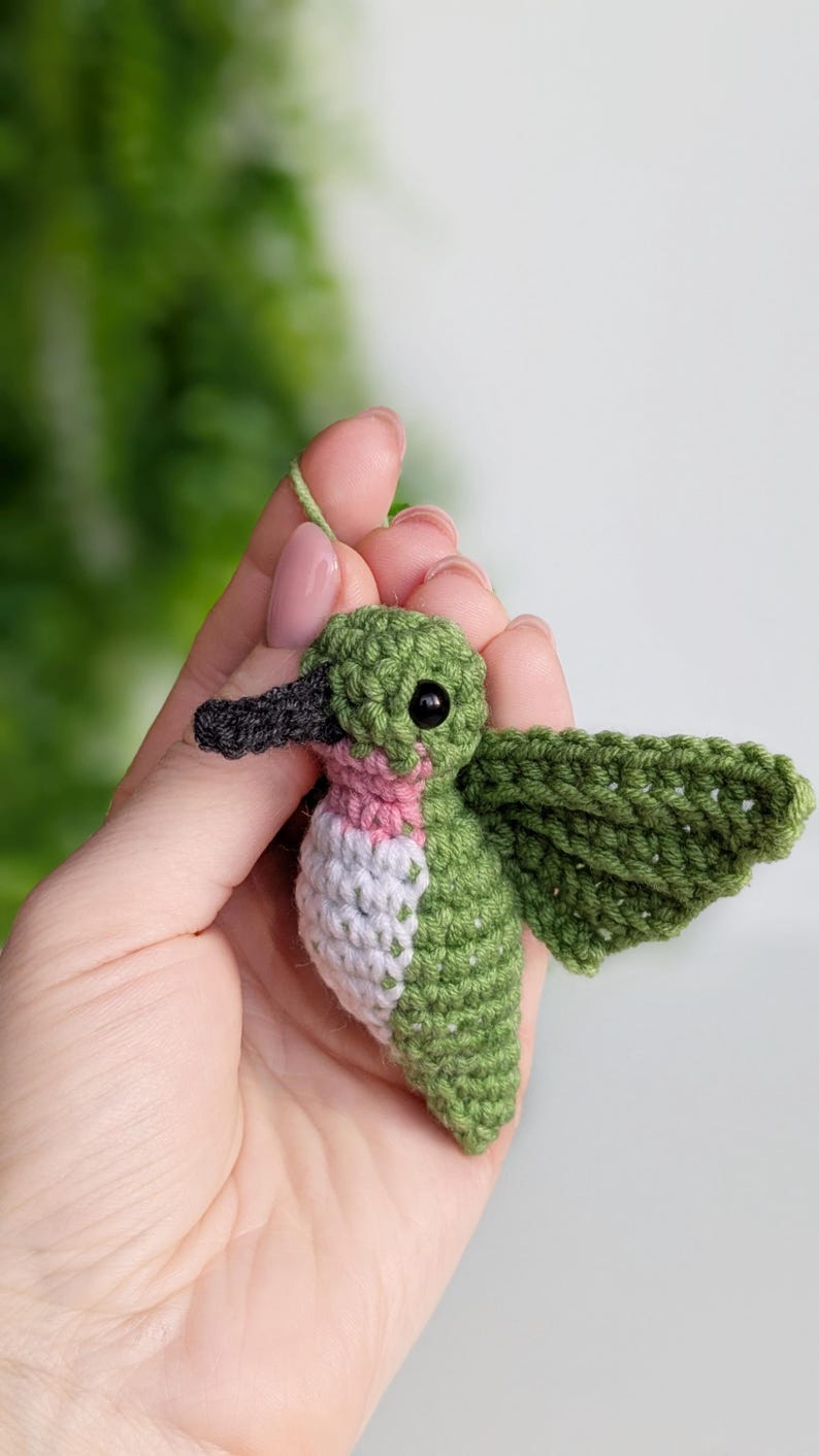 May include: A small green and pink crocheted hummingbird with black eyes and a black beak. The hummingbird is being held in a hand.