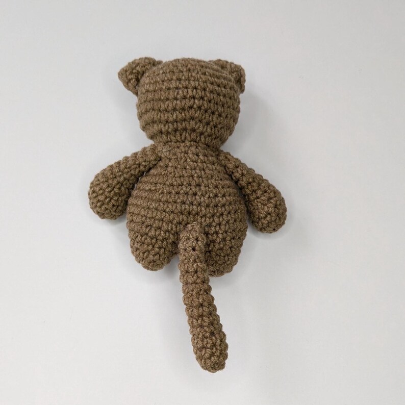 May include: A brown crocheted stuffed animal featuring a rounded head with two small ears, a plump body, two short arms, and a long tail extending downward. The toy is crafted from yarn with a visible textured stitch pattern throughout the entire form.