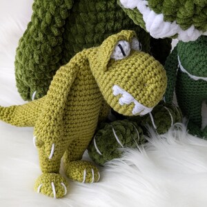 Puede incluir: Juguetes de dinosaurios de ganchillo hechos a mano en varios tonos de verde. El dinosaurio principal es de color verde claro con detalles blancos en los dientes y las garras. El fondo presenta un dinosaurio m&aacute;s grande, de color verde oscuro y una superficie blanca y esponjosa.