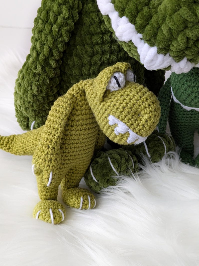 Puede incluir: Juguetes de dinosaurios de ganchillo hechos a mano en varios tonos de verde. El dinosaurio principal es de color verde claro con detalles blancos en los dientes y las garras. El fondo presenta un dinosaurio m&aacute;s grande, de color verde oscuro y una superficie blanca y esponjosa.