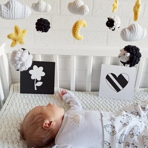 May include: A white crib with a black and white mobile hanging above a baby sleeping on a white blanket with a sheep pattern. The mobile features crocheted clouds, stars, a sun, a moon, and sheep.