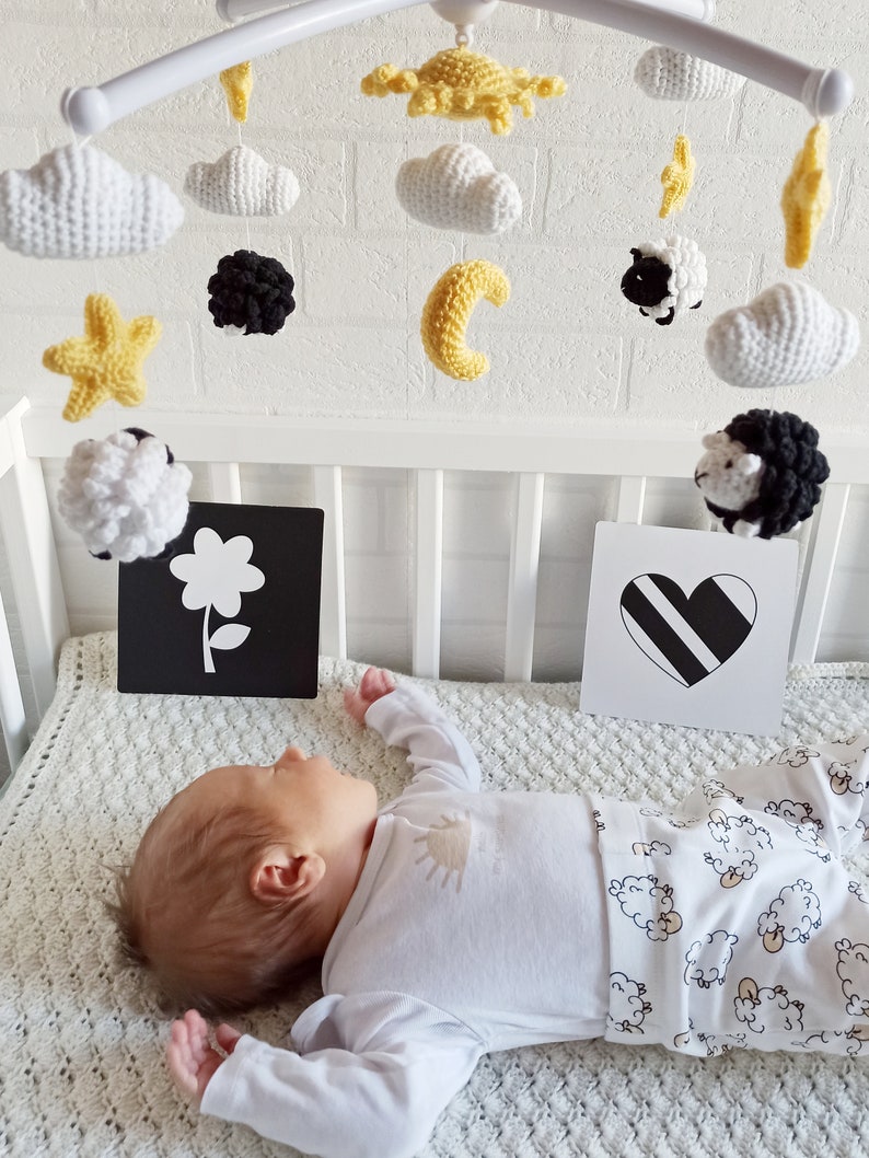 May include: A white crib with a black and white mobile hanging above a baby sleeping on a white blanket with a sheep pattern. The mobile features crocheted clouds, stars, a sun, a moon, and sheep.