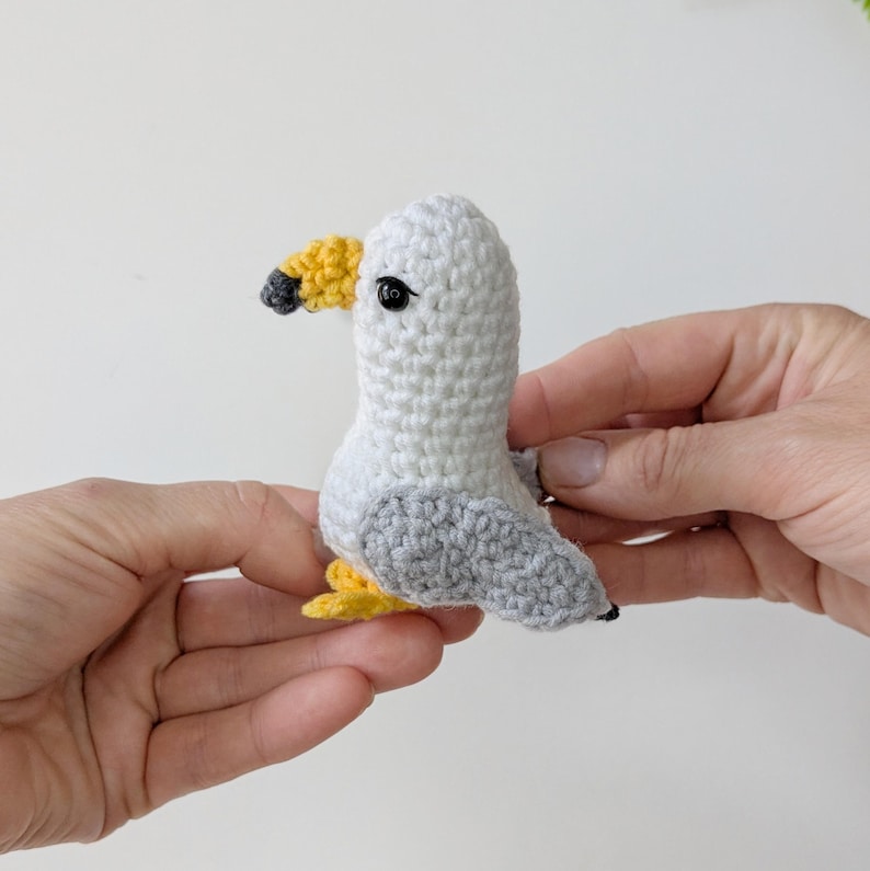 Op de afbeelding: Een klein, handgemaakt gehaakt meeuwspeeltje met een witte kop en lichaam, lichtgrijze vleugels, een gele snavel met een donkergrijze punt en kleine gele pootjes. De vogel heeft &eacute;&eacute;n zwart plastic kraaloog aan de zijkant van de kop.