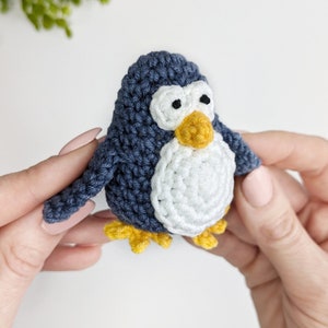 Pode incluir: Um brinquedo de pinguim de croch&ecirc; com corpo azul e branco, bico e p&eacute;s amarelos. O pinguim est&aacute; sendo segurado por duas m&atilde;os.