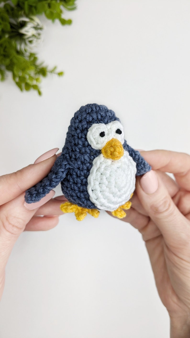 Pode incluir: Um brinquedo de pinguim de croch&ecirc; com corpo azul e branco, bico e p&eacute;s amarelos. O pinguim est&aacute; sendo segurado por duas m&atilde;os.