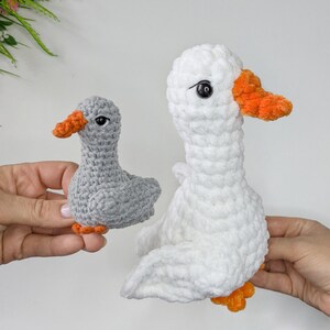 May include: Two handmade crochet plush toys shaped like geese. One goose is smaller and light gray with an orange beak and feet. The other goose is larger, made of white plush yarn, and features a bright orange beak and orange feet with black eyes.