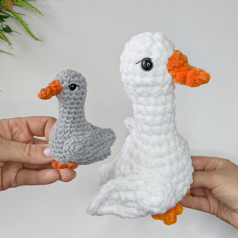 May include: Two handmade crochet plush toys shaped like geese. One goose is smaller and light gray with an orange beak and feet. The other goose is larger, made of white plush yarn, and features a bright orange beak and orange feet with black eyes.