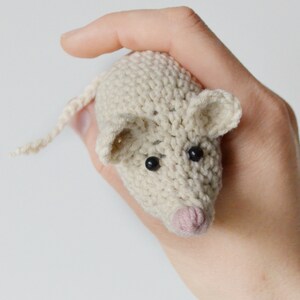 May include: A crocheted white mouse toy with black eyes and a pink nose. The mouse is being held by a hand.