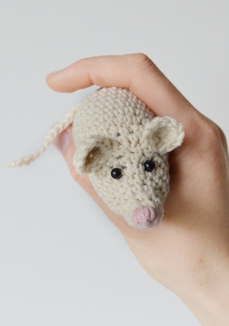 May include: A crocheted white mouse toy with black eyes and a pink nose. The mouse is being held by a hand.
