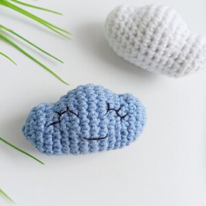 May include: Two crocheted clouds, one blue with a smiling face and one white. The clouds are on a white background.