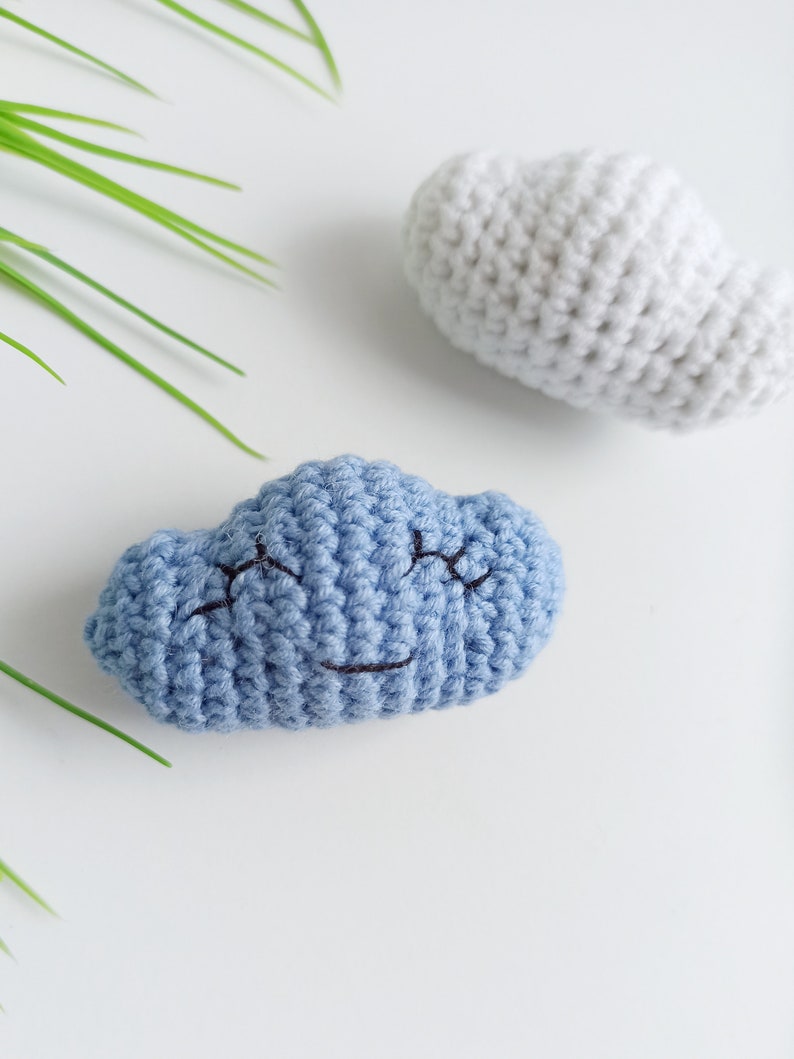 May include: Two crocheted clouds, one blue with a smiling face and one white. The clouds are on a white background.