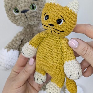 K&ouml;nnte beinhalten: Zwei handgefertigte H&auml;keltierspielzeuge in Katzenform. Eines ist senfgelb mit wei&szlig;en Akzenten an Ohren und Pfoten. Das andere ist eine hellbraune Katze mit wei&szlig;en Akzenten. Beide haben schwarze Schnurrhaare und gr&uuml;ne oder schwarze Augen.