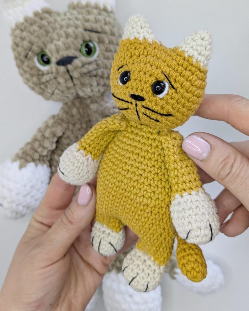K&ouml;nnte beinhalten: Zwei handgefertigte H&auml;keltierspielzeuge in Katzenform. Eines ist senfgelb mit wei&szlig;en Akzenten an Ohren und Pfoten. Das andere ist eine hellbraune Katze mit wei&szlig;en Akzenten. Beide haben schwarze Schnurrhaare und gr&uuml;ne oder schwarze Augen.