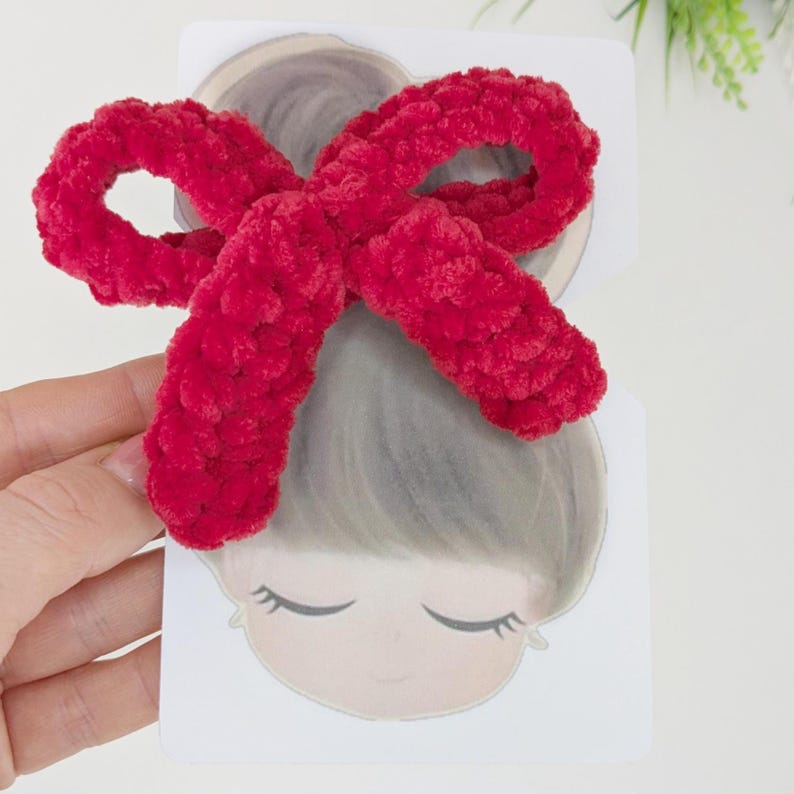 May include: A bright red, fluffy bow hair clip. The bow is made of a soft, textured material and is positioned over a cartoon illustration of a person with closed eyes. The clip is likely for hair styling.