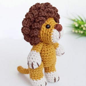 May include: A crocheted lion toy with a brown mane, yellow body, and white paws. The lion is standing on a white surface.