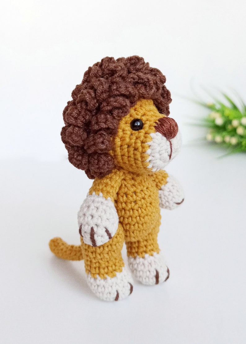May include: A crocheted lion toy with a brown mane, yellow body, and white paws. The lion is standing on a white surface.