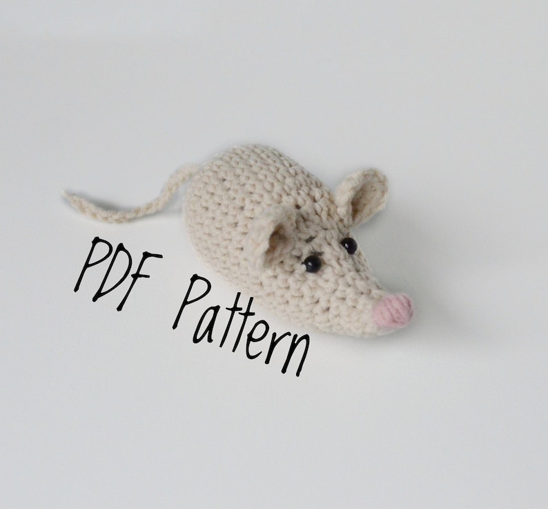 May include: A crocheted mouse amigurumi toy with black eyes and a pink nose. The toy is laying on a white surface with the text "PDF Pattern" written below it.