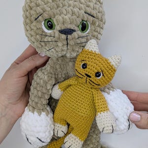 K&ouml;nnte beinhalten: Zwei Pl&uuml;schkatzenspielzeuge. Die gr&ouml;&szlig;ere Katze ist hellbraun mit wei&szlig;en Akzenten an Ohren und Pfoten und gr&uuml;nen Augen. Ein kleineres, gelbes Katzenspielzeug mit wei&szlig;en Akzenten wird vor der gr&ouml;&szlig;eren gehalten. Beide sind handgefertigt.