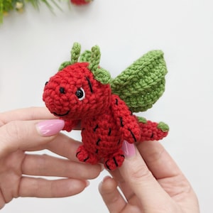 May include: A small, red crocheted dragon with green wings. The dragon has black eyes and a smiling mouth.
