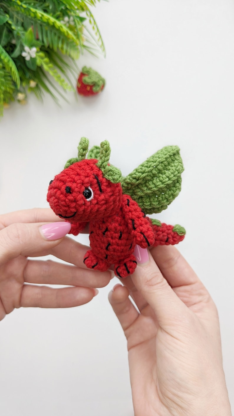 May include: A small, red crocheted dragon with green wings. The dragon has black eyes and a smiling mouth.