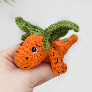 May include: A crocheted carrot-shaped toy with green leaves for wings. The toy has black eyes and a smiling mouth.