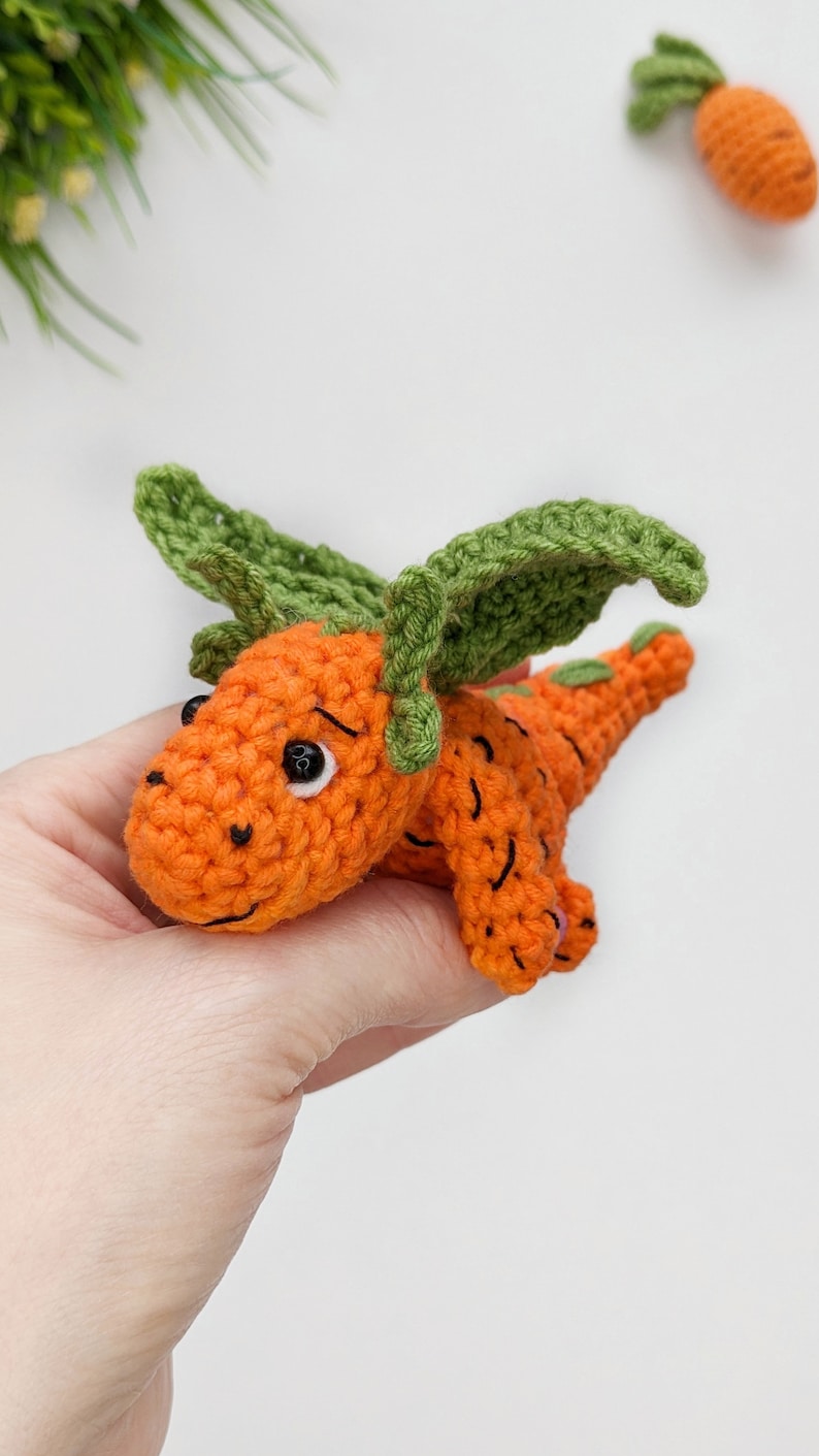May include: A crocheted carrot-shaped toy with green leaves for wings. The toy has black eyes and a smiling mouth.