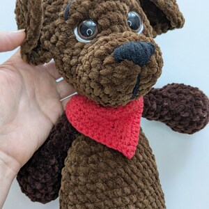 May include: A brown crocheted plush dog toy featuring a textured yarn finish, large dark plastic eyes, a black stitched nose, and a bright red triangular bandana tied around its neck. The stuffed animal has floppy ears and a soft, rounded body.
