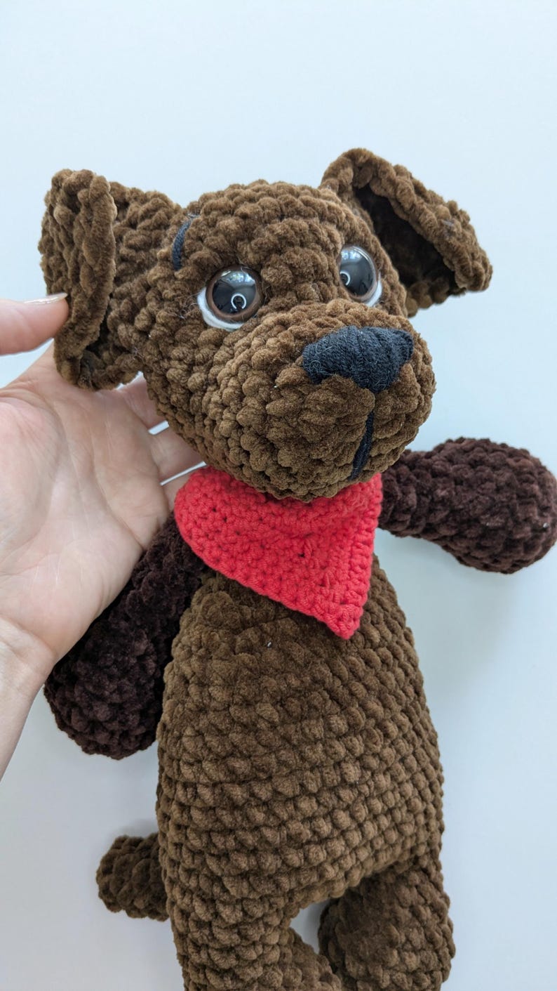 May include: A brown crocheted plush dog toy featuring a textured yarn finish, large dark plastic eyes, a black stitched nose, and a bright red triangular bandana tied around its neck. The stuffed animal has floppy ears and a soft, rounded body.