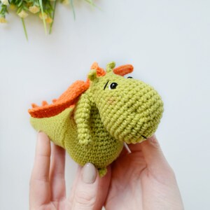 May include: A green crocheted dragon toy with orange wings and black eyes. The dragon is being held by two hands.