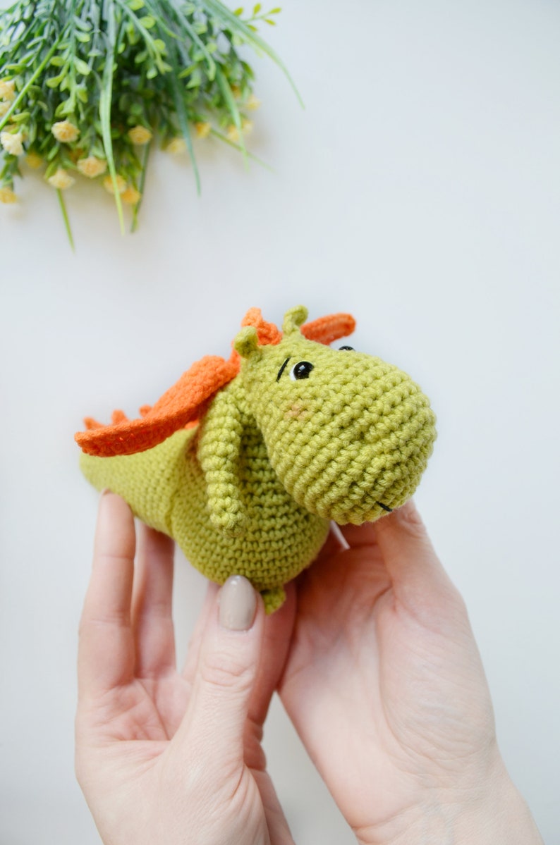 May include: A green crocheted dragon toy with orange wings and black eyes. The dragon is being held by two hands.